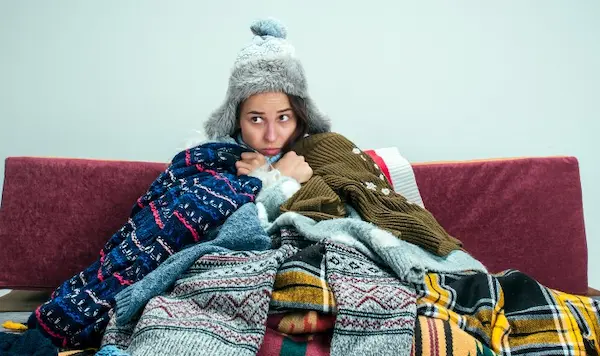 Mujer con gorro y bufanda envuelta en varias mantas en un sofá para combatir el frío extremo.